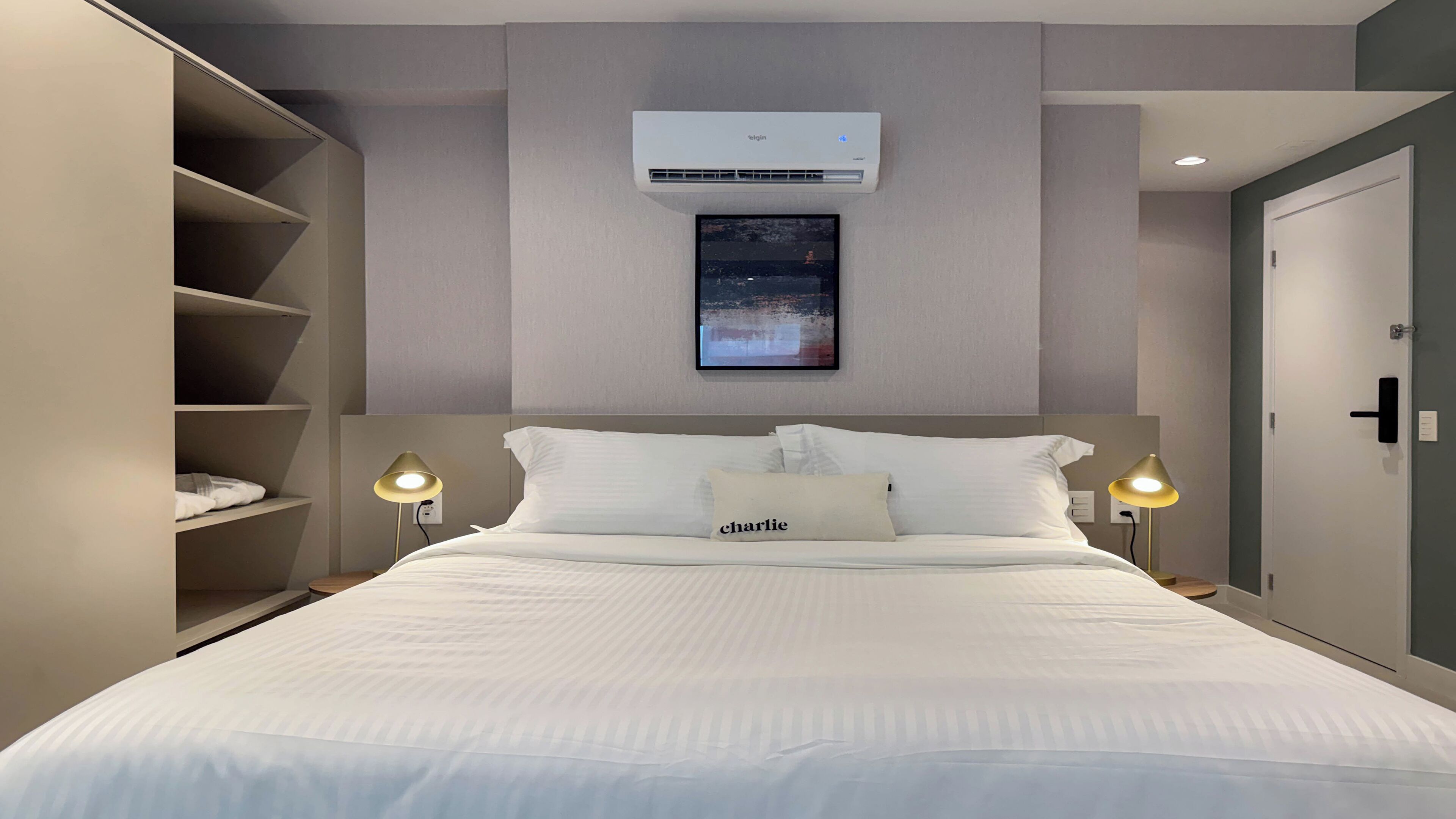 Un lit bien fait, avec une literie blanche et un oreiller sur lequel est écrit le mot « Charlie ». Un climatiseur mural est fixé au-dessus du lit, et un tableau encadré est accroché au mur.