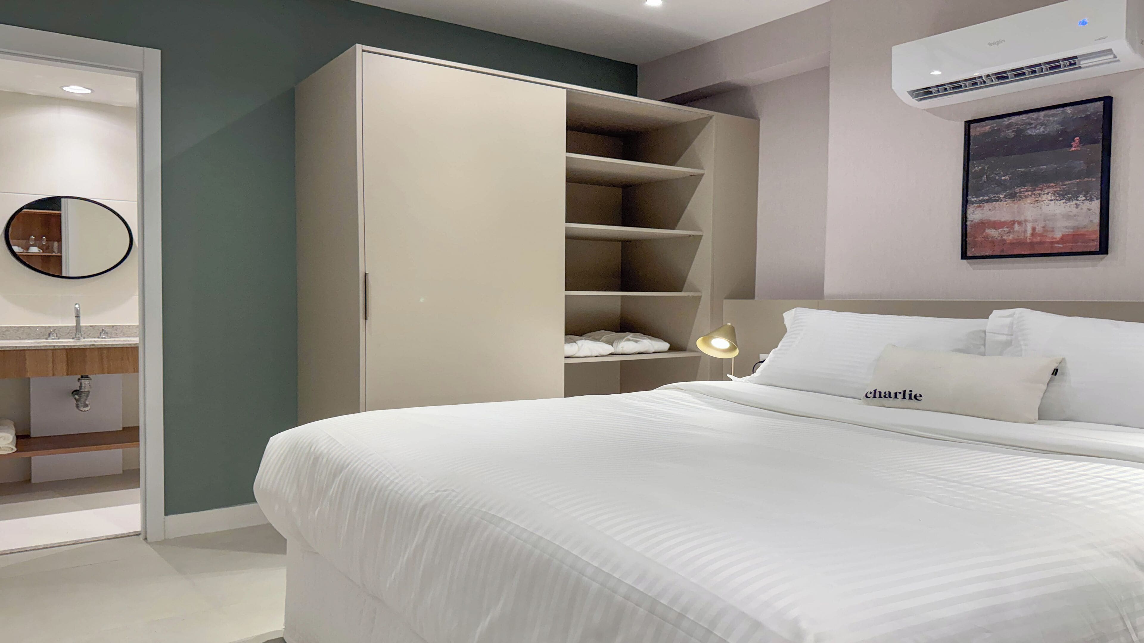 A bedroom with a bed, a wardrobe, a bedside table with a lamp, a mirror, and a wall-mounted air conditioner.