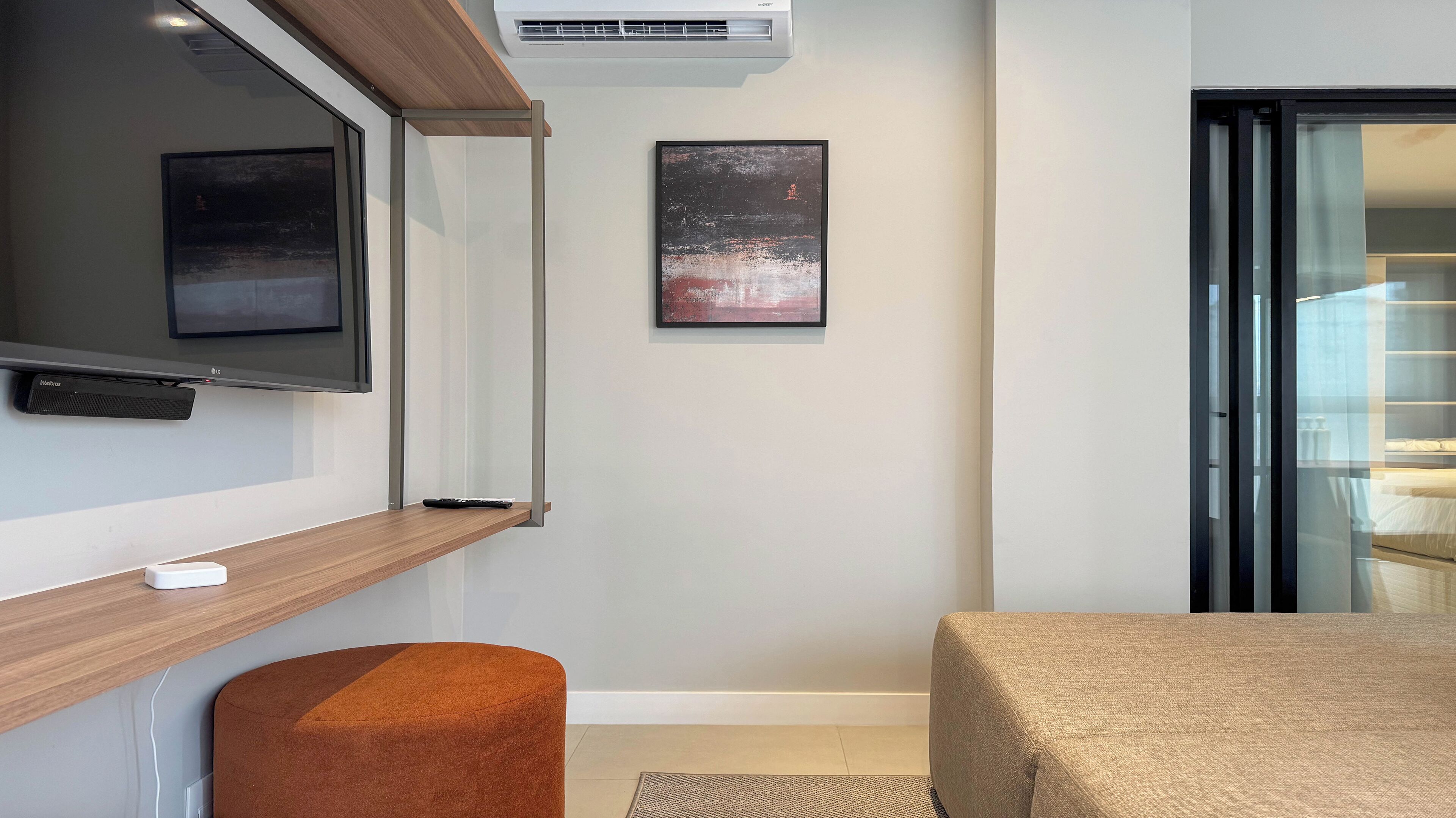 A modern hotel room with a flat-screen TV mounted on the wall, a small bedside table, a single bed, and a sliding door leading to a balcony.