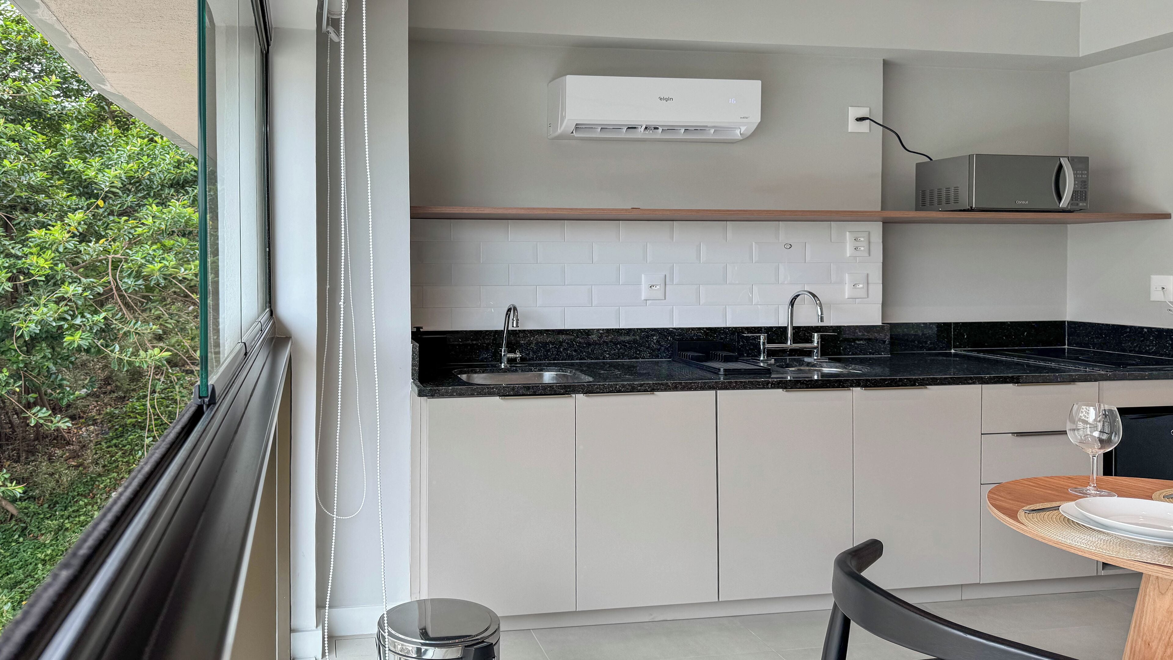 A modern kitchen with a black countertop, white cabinets, a built-in oven, and a window overlooking greenery.