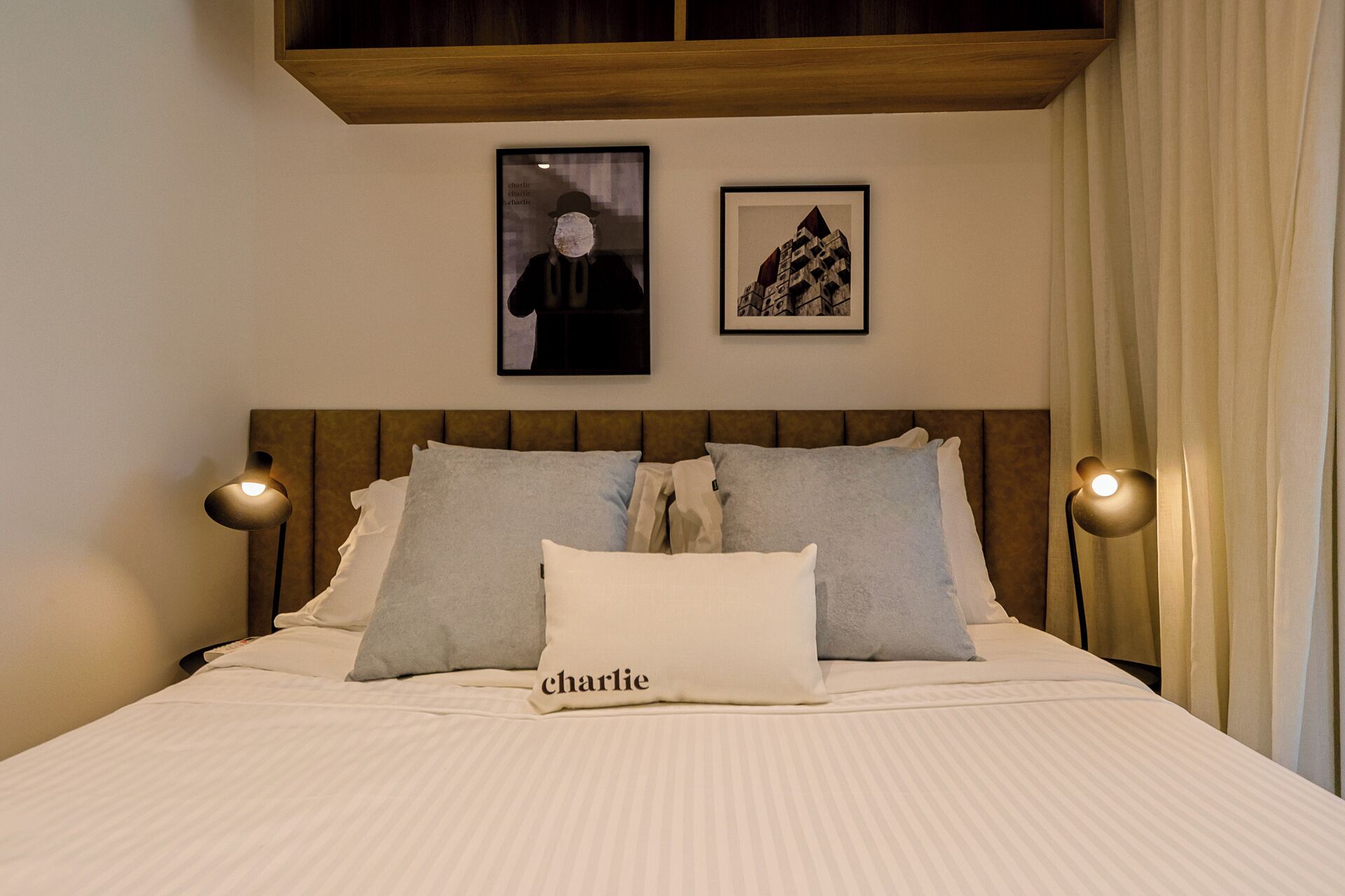 A neatly made bed with a pillow labeled "charlie" and two wall-mounted lamps.