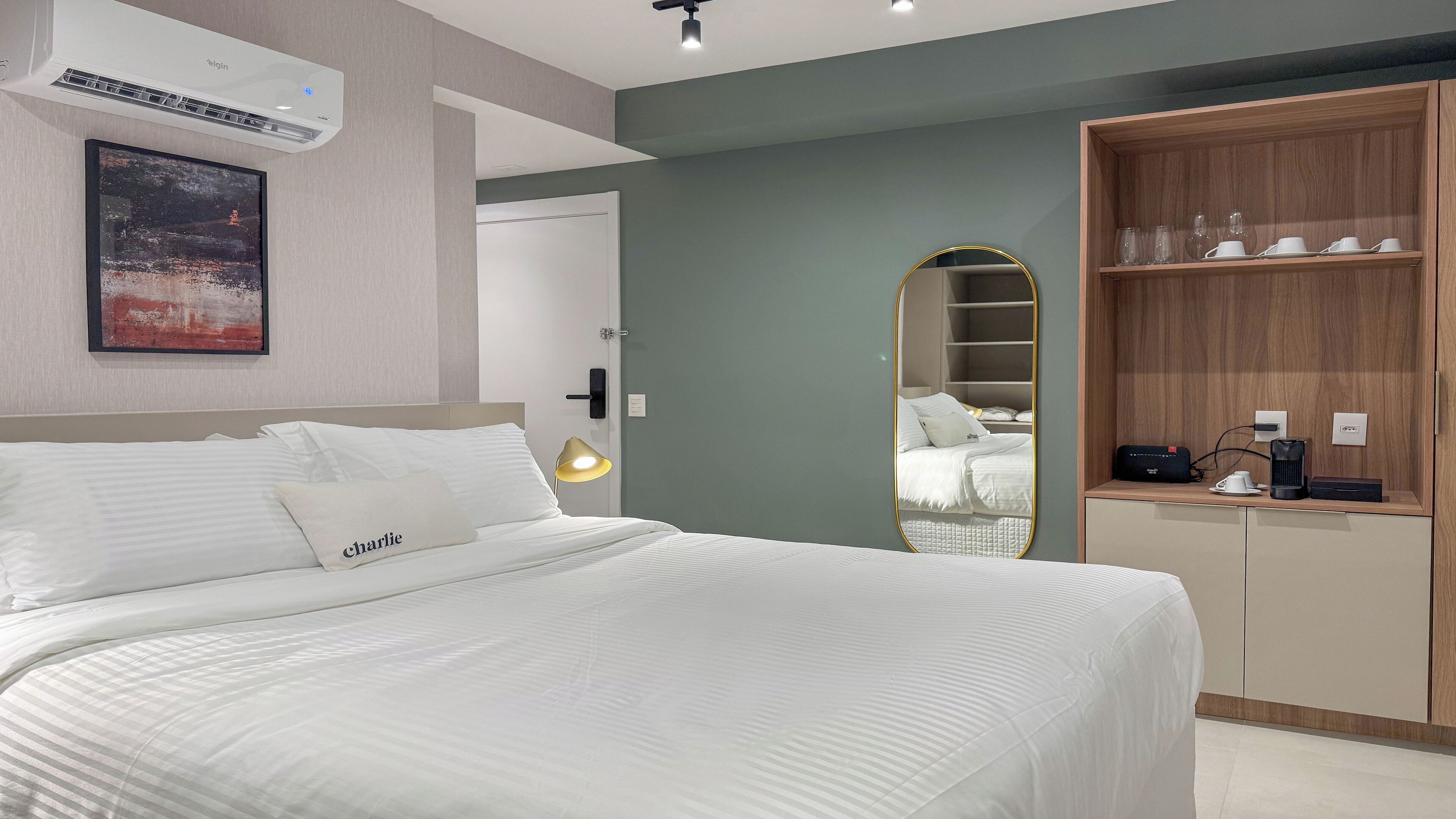 A modern hotel room with a neatly made bed, a bedside lamp, a large mirror, and a wooden wardrobe with shelves.