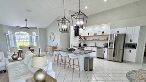 Amplia sala de estar con un sofá grande, una mesa de comedor con taburetes tipo bar, una cocina con electrodomésticos modernos y un ventilador de techo.
