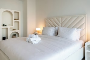 A neatly made bed with white linens and rolled towels, a headboard with a geometric pattern, and a bedside table with a lamp and decorative items. - Pure Voyage Beachfront  (Dubai)