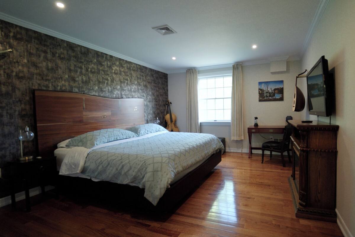 A bedroom with a large bed, a wooden desk, a chair, a guitar, and a television.