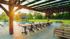 Un espace extérieur avec une piscine, une longue table en bois et des chaises, le tout entouré d’une terrasse couverte illuminée de guirlandes lumineuses.