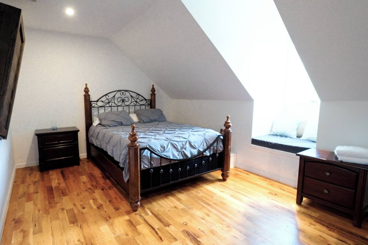 A bedroom with a wooden floor, a bed with a headboard, a nightstand, a dresser, and a window with a view.