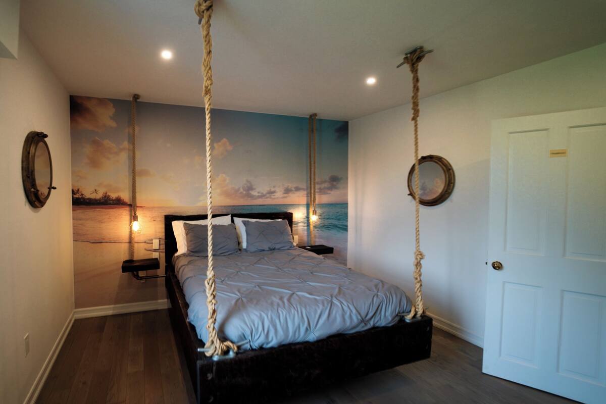 A bedroom with a bed suspended from ropes, a mural of a sunset over water, and a door with a nameplate.