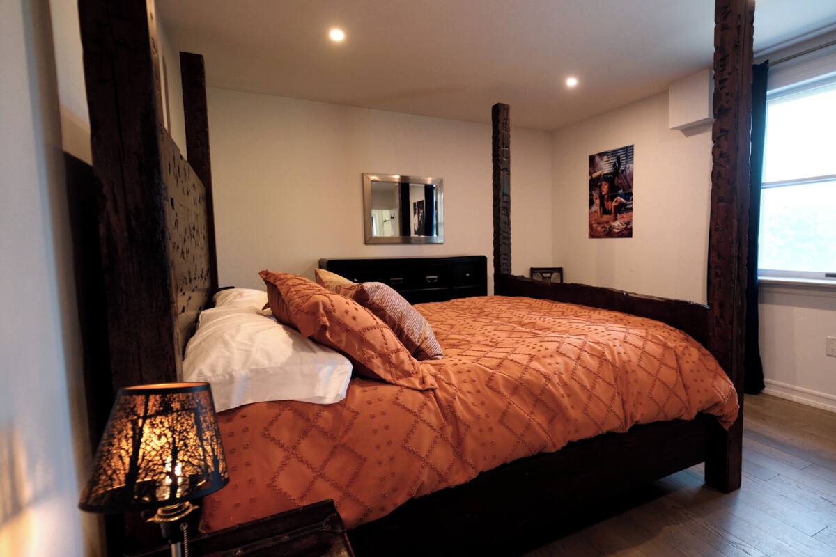 A bedroom with a large bed, a bedside table, a lamp, a mirror, and a window with curtains.