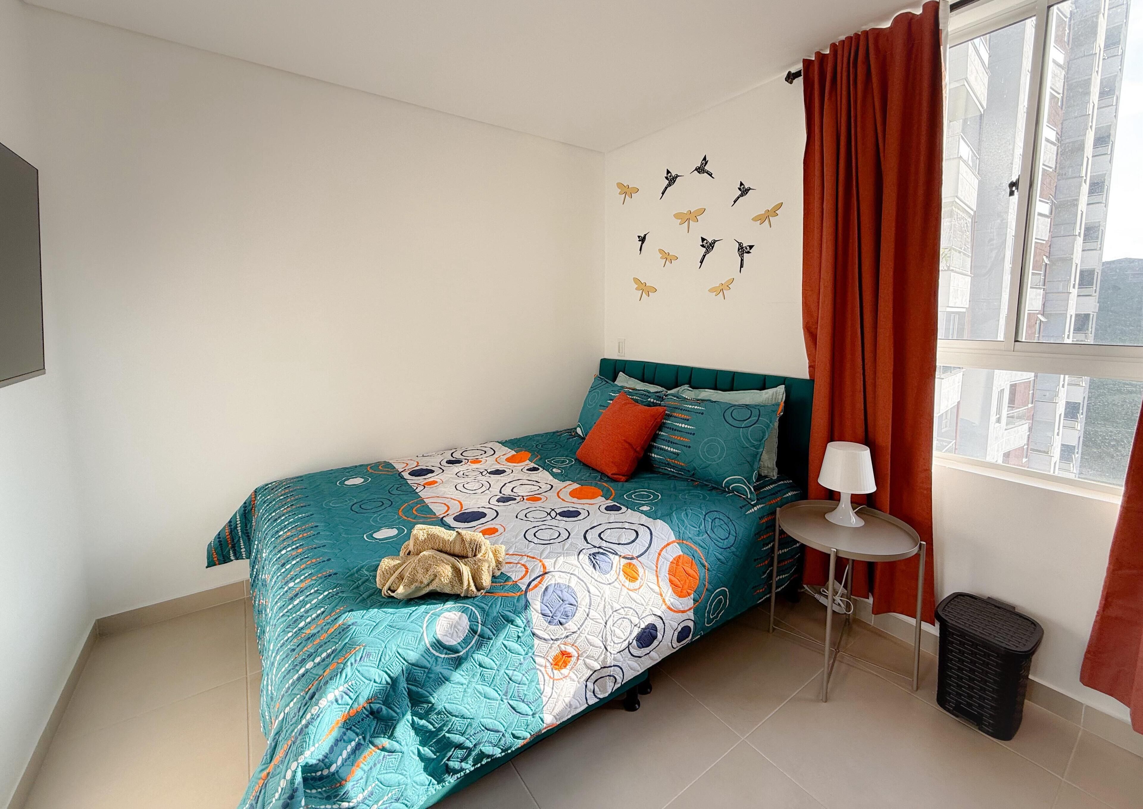 A bedroom with a bed, a nightstand, a lamp, a window with curtains, and a wall decal of birds.