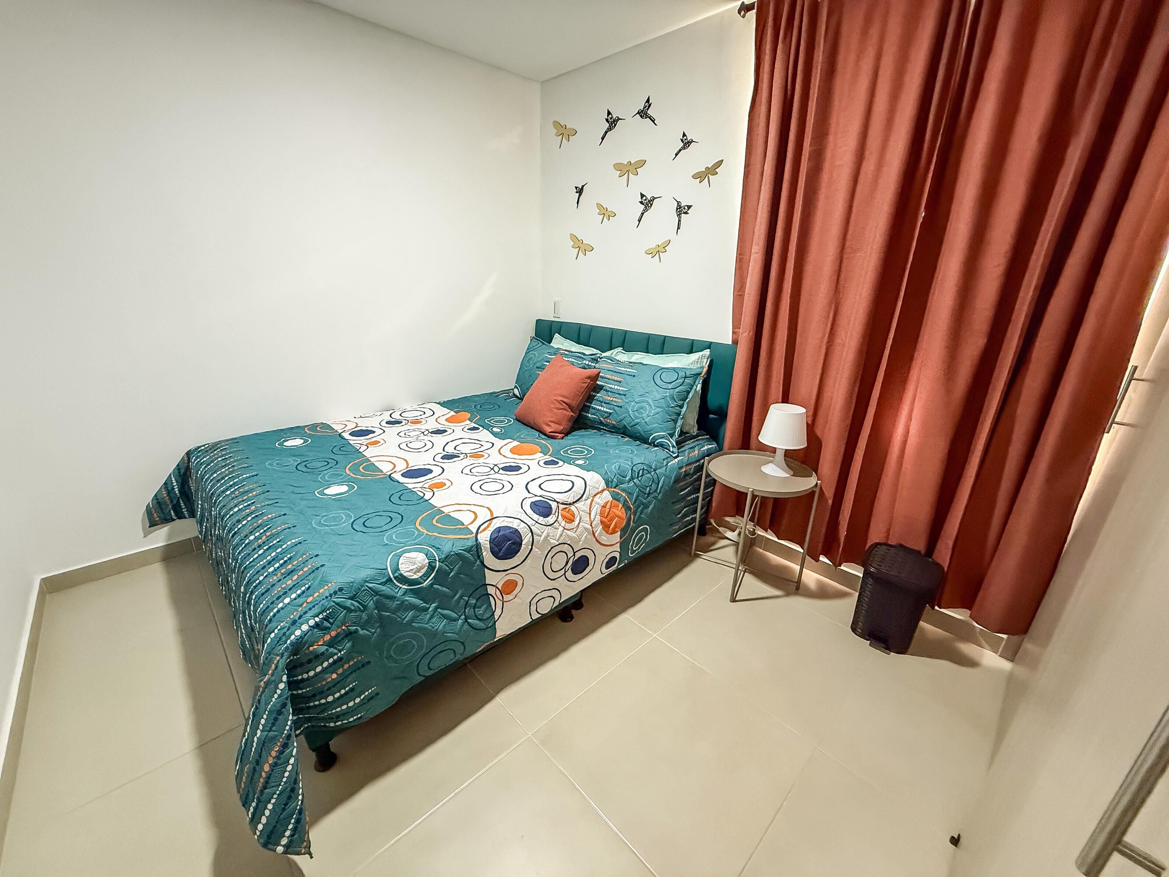 Un dormitorio con cama, almohadas, una mesita de noche, una lámpara y una cortina.