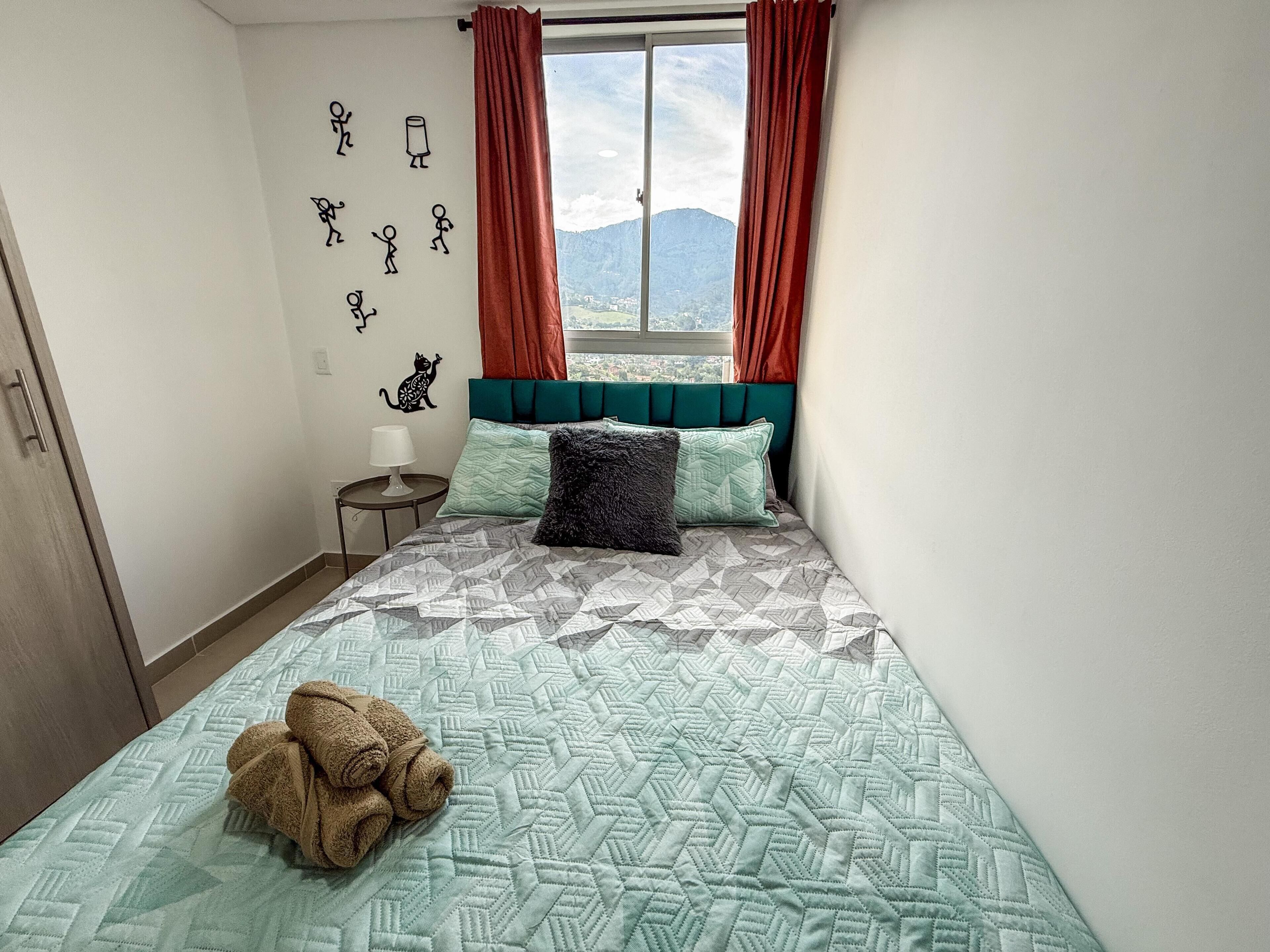 A bedroom with a bed, a window with curtains, a wall with stick figure drawings, and a towel on the bed.
