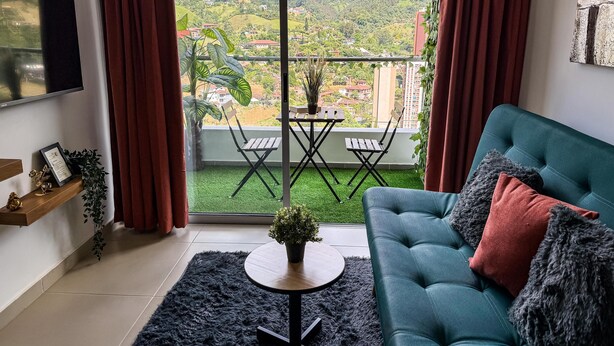 A living room with a sofa, a small table, and a view of a green hill and buildings. - Mountain View, Pool & Gym (2506) (Sabaneta)