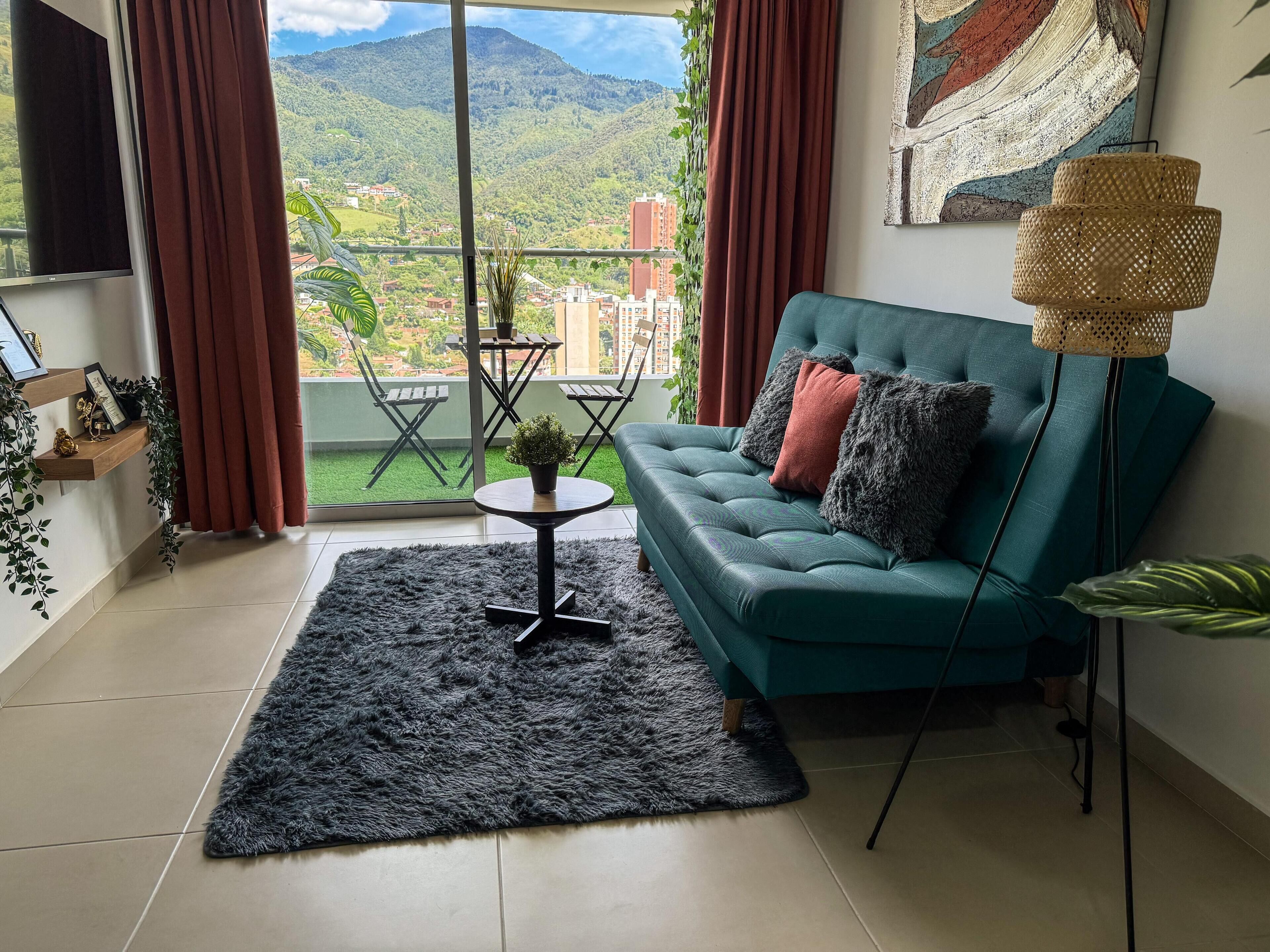 Ein Wohnzimmer mit einem türkisfarbenen Sofa, einem kleinen runden Tisch und Blick auf eine grüne Landschaft durch große Fenster.