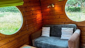 L’intérieur chaleureux d’un chalet avec un banc rembourré, une petite table en bois et deux fenêtres rondes donnant sur un paysage verdoyant.