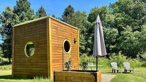 Une cabane en bois dotée de deux fenêtres rondes, d’un jardin surélevé planté de végétaux et d’un espace extérieur aménagé avec des chaises et un parasol.