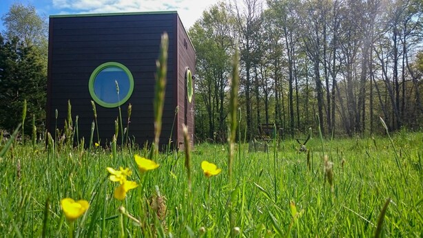 Un bâtiment moderne doté d’une fenêtre circulaire et d’une porte verte, entouré d’un champ verdoyant et d’arbres.