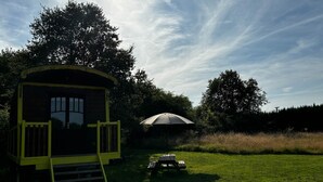 Una pequeña estructura de madera con una puerta amarilla y escalones, una mesa de picnic con sombrilla, y un campo cubierto de césped.