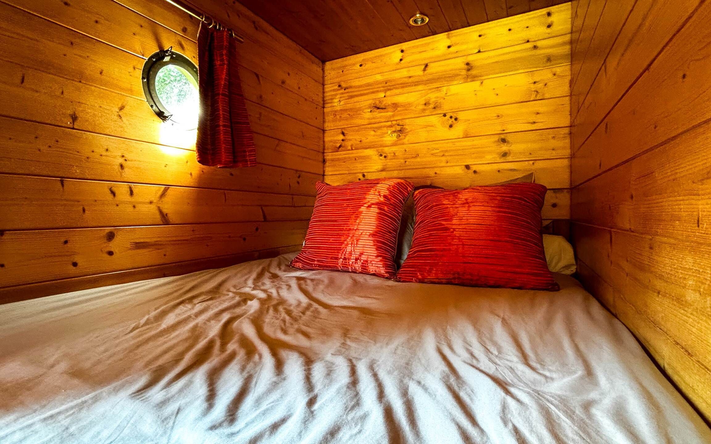 A wooden cabin room with a bed, red pillows, and a window with curtains.