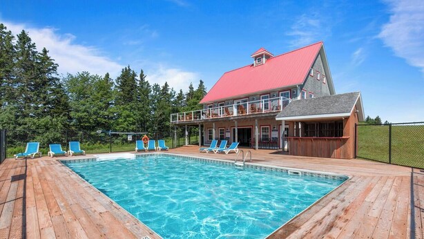 Ein Schwimmbecken mit Holzterrasse und Liegestühlen, ein Gebäude mit rotem Dach sowie ein von Bäumen gesäumtes, eingezäuntes Areal.