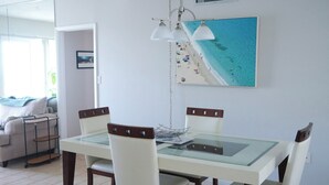 A dining area with a glass-top table, four chairs, a hanging light fixture, and a beach-themed painting on the wall. - Sunny Escape 2-Bedroom Condo Steps from Cocoa Beach (Cocoa Beach)