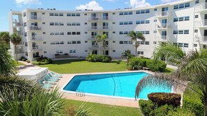 A multi-story hotel with a swimming pool surrounded by greenery and palm trees. - Sunny Escape 2-Bedroom Condo Steps from Cocoa Beach (Cocoa Beach)