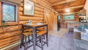 A wooden cabin interior with a dining area, kitchen, and a large window overlooking greenery. - Ski, Hike & Snowshoe: Homer Getaway w/ Fire Pit (Homer)