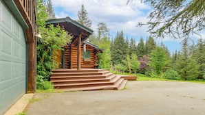 A log cabin with a green garage door, wooden steps leading to a porch, and a forested backdrop. - Ski, Hike & Snowshoe: Homer Getaway w/ Fire Pit (Homer)
