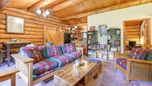A cozy log cabin interior with a wooden ceiling, a colorful sofa, a wooden coffee table, and a bookshelf filled with books and decorative items. - Ski, Hike & Snowshoe: Homer Getaway w/ Fire Pit (Homer)