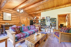 Interior de una cabaña de troncos con techo de madera, un sofá de colores, una mesa de centro de madera y una zona de cocina con armarios de madera y un refrigerador.