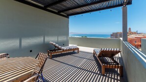 A balcony with a view of the ocean and buildings. There are two wooden lounge chairs and a table.