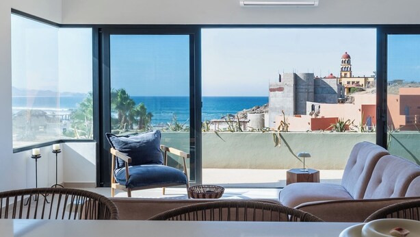 A modern living room with a large window overlooking a coastal view, including a beach and buildings. - Pico del Punto • New Penthouse with Panoramic Views, Surf Access & Rooftop Deck (Los Cerritos)
