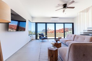 Un salon moderne doté d’une grande fenêtre, d’un ventilateur de plafond, d’un canapé, d’un fauteuil et d’un téléviseur fixé au mur.
