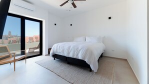A modern bedroom with a large bed, a ceiling fan, a view of the city through a large window, and a balcony with a chair. - Pico del Punto • New Penthouse with Panoramic Views, Surf Access & Rooftop Deck (Los Cerritos)