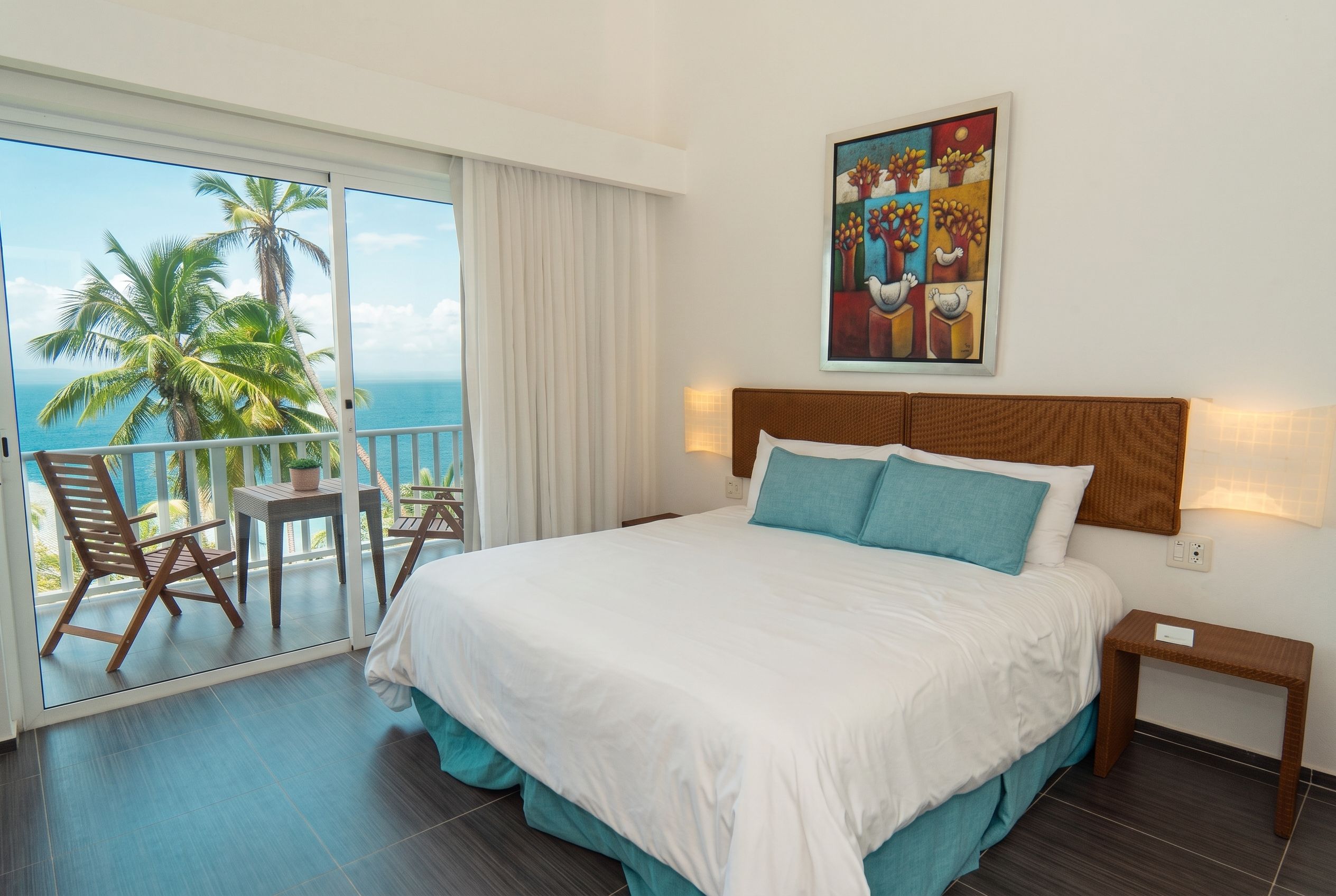 Habitación de hotel con una cama grande, un balcón con vista a palmeras y al mar, y un cuadro en la pared.