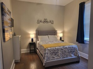 Quarto com cama, duas mesas de cabeceira, um criado-mudo, um objeto decorativo fixado na parede, uma janela com persianas e um quadro na parede.