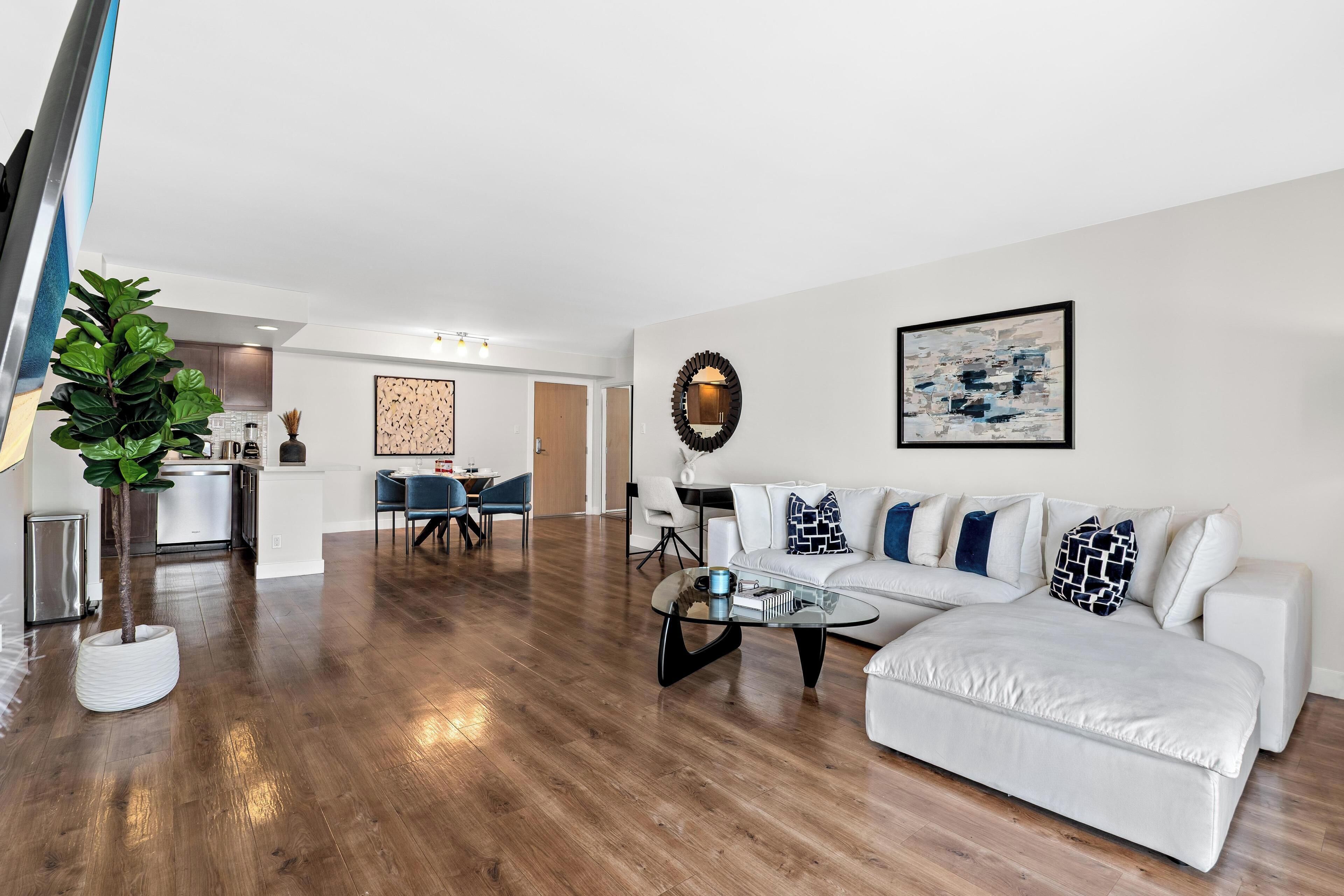 A spacious living room with a white sofa, a glass coffee table, and a dining area in the background.