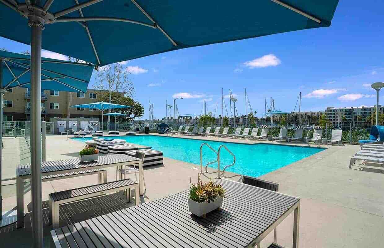 A pool area with lounge chairs and umbrellas, surrounded by buildings and a clear sky.