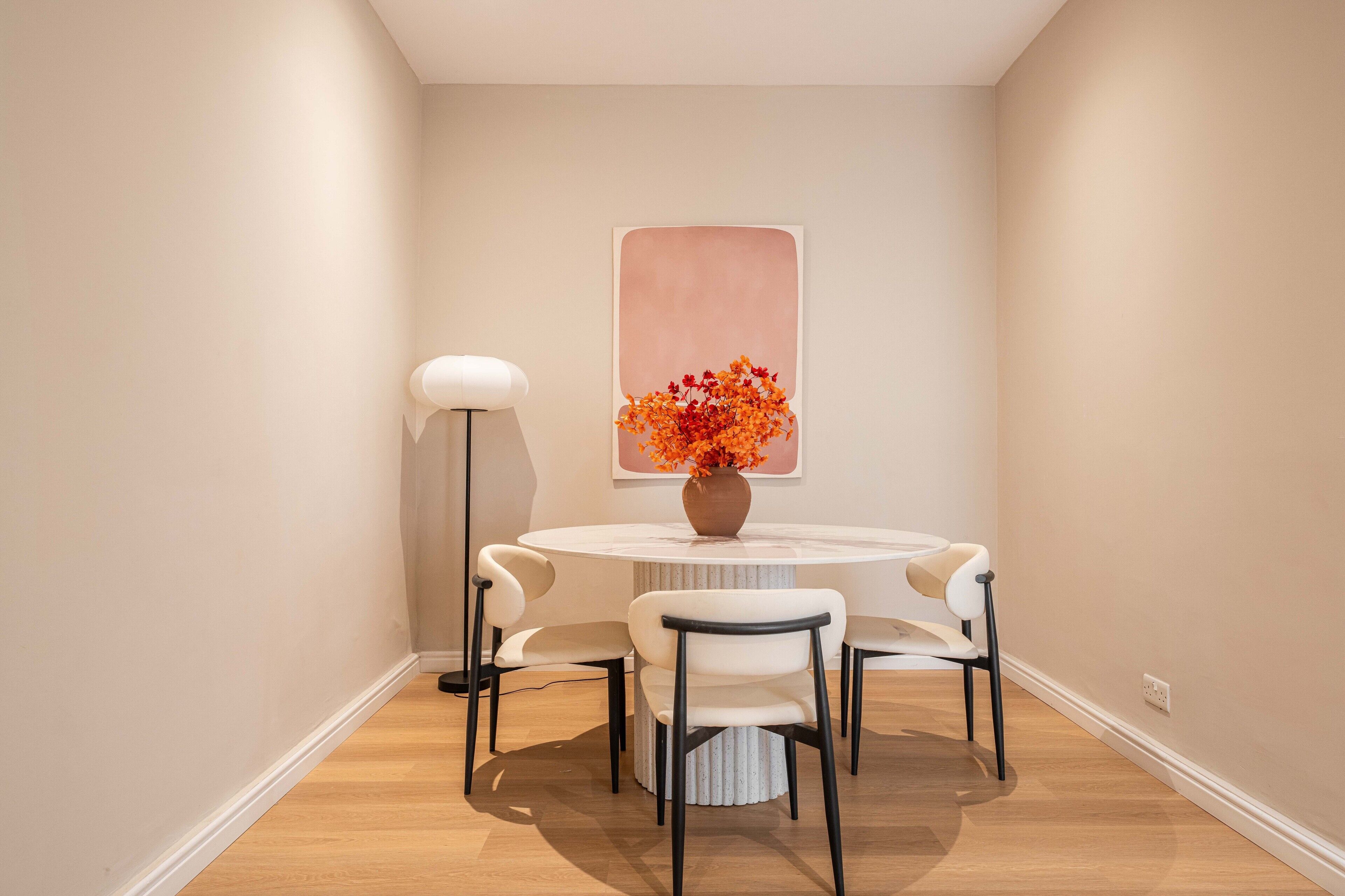 A dining area with a round table, four chairs, a vase with flowers, a floor lamp, and a large abstract painting on the wall.