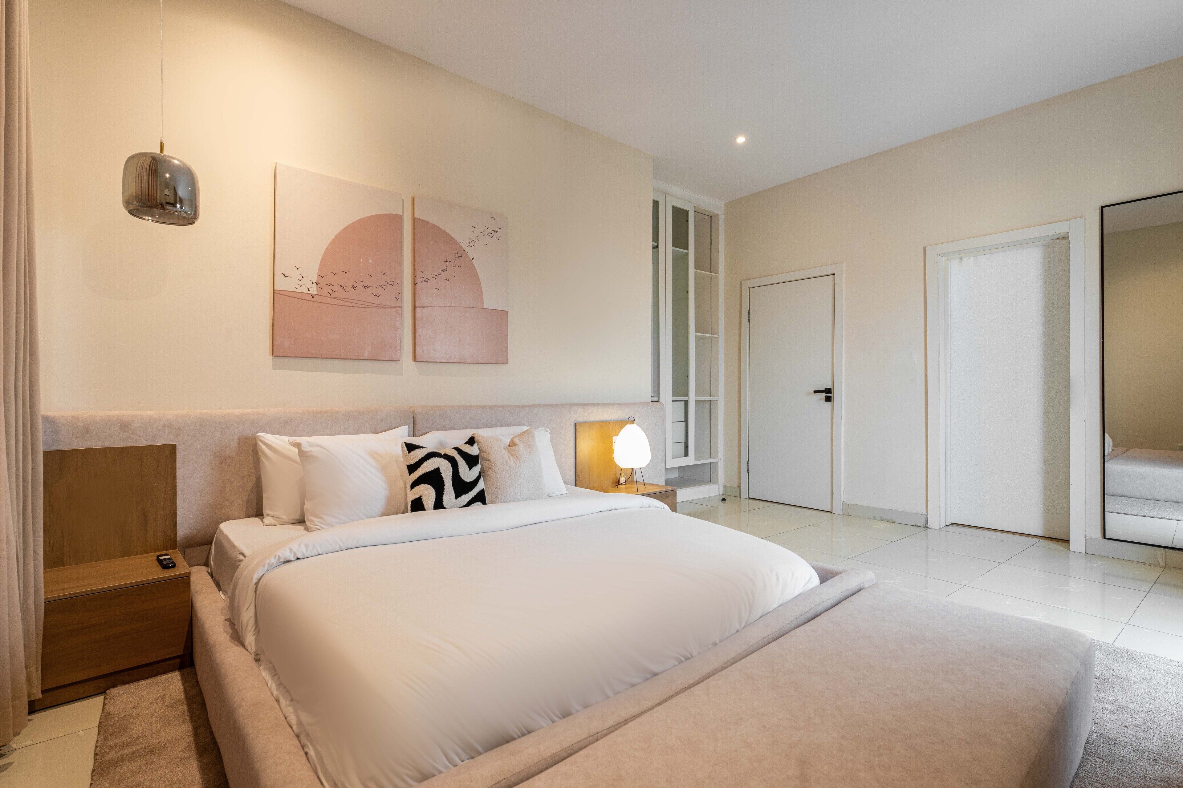 A modern bedroom with a large bed, bedside tables, a wall-mounted lamp, and a door leading to another room.