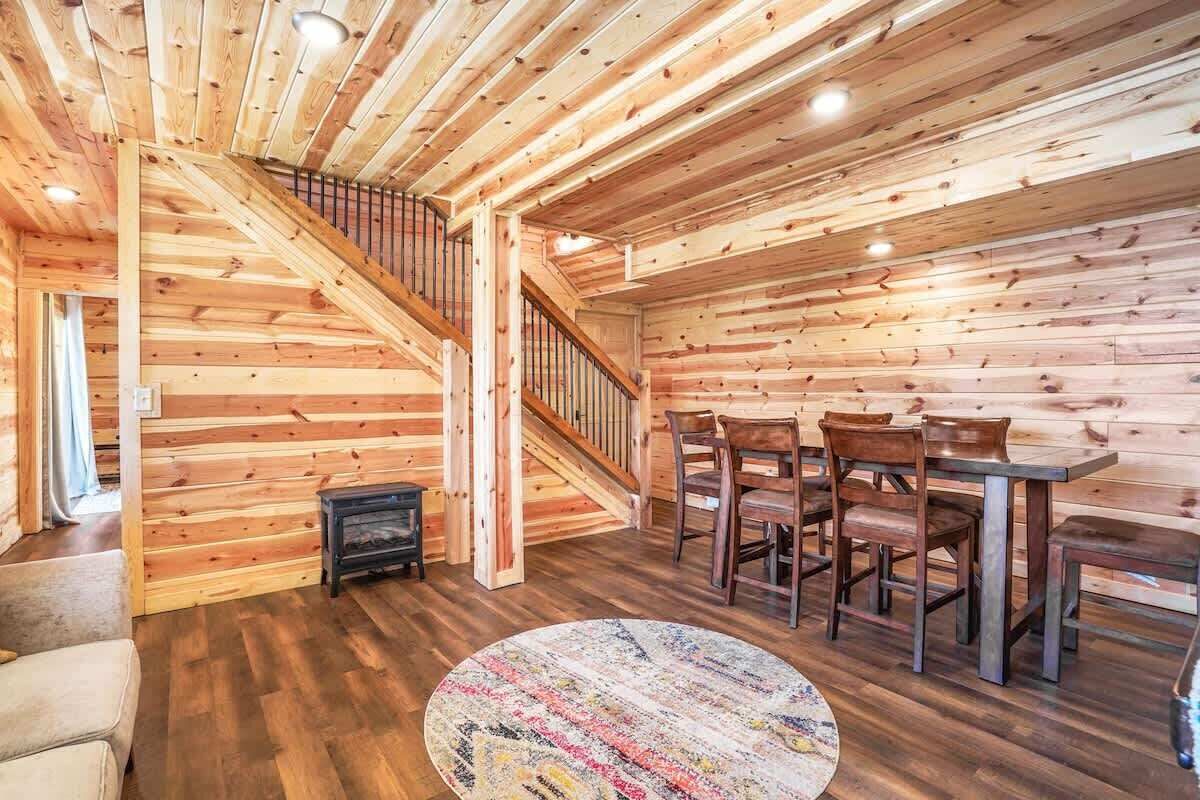 A wooden interior with a staircase, dining area, and a round rug.