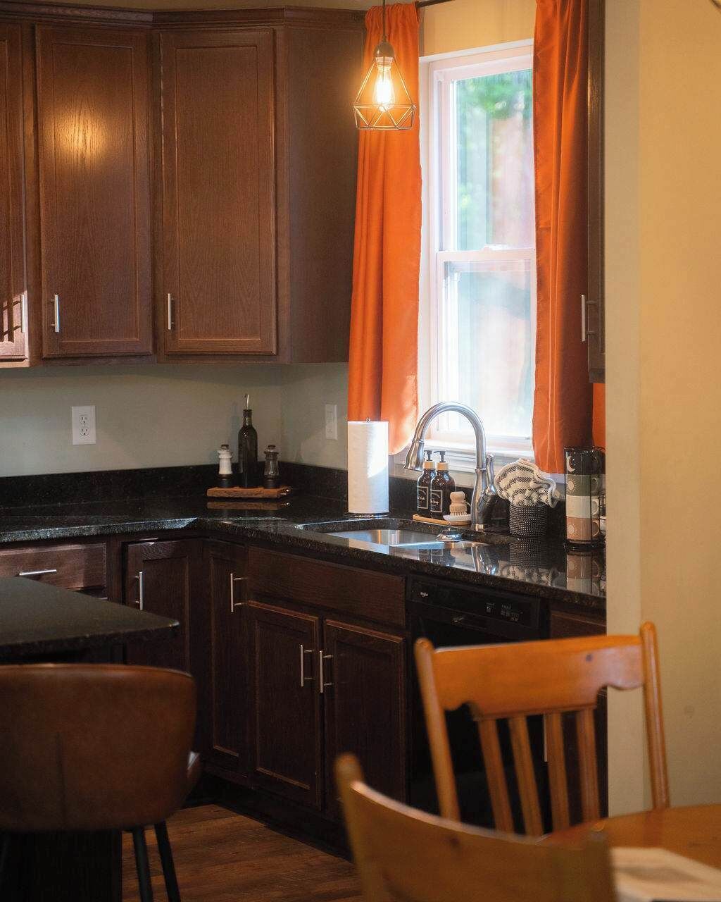 Una cocina con armarios de madera, encimera negra, un lavamanos y una ventana con cortinas naranjas.