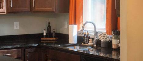 Una cocina con armarios de madera, encimera negra, un lavamanos y una ventana con cortinas naranjas.