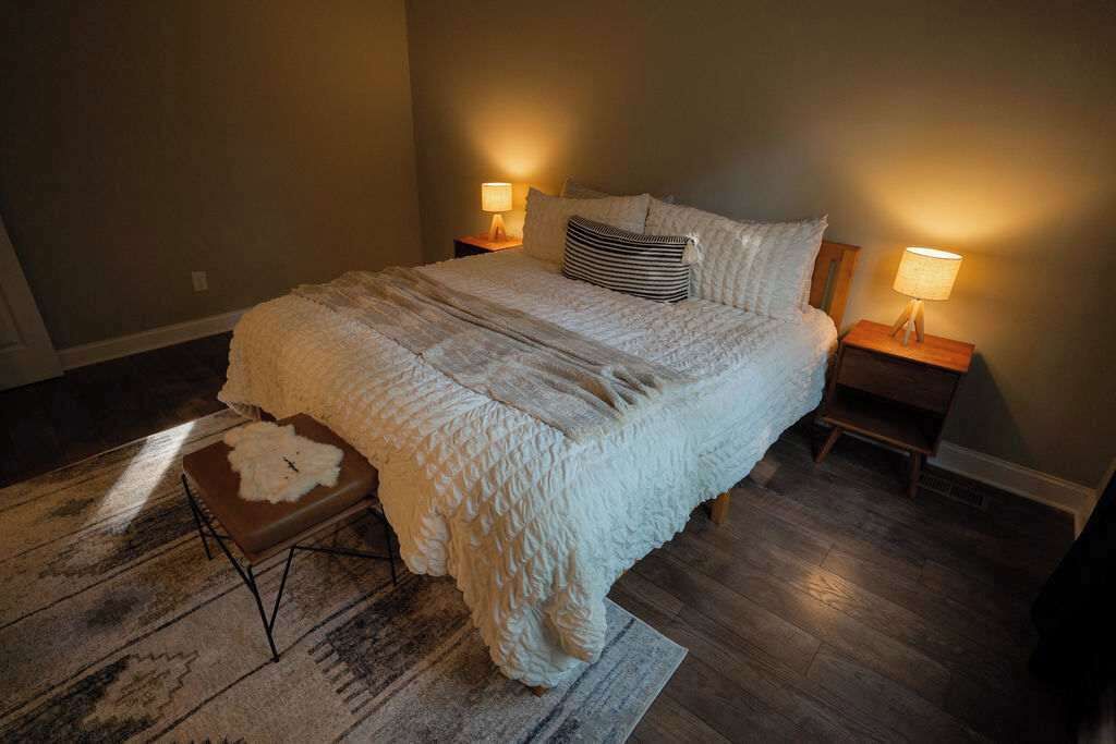 Un dormitorio con una cama grande, dos lámparas de noche, una alfombra y un taburete pequeño.