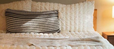 Una cama bien hecha con una almohada a rayas y una colcha blanca, una mesita de noche de madera con una lámpara, y una alfombra estampada en el suelo.