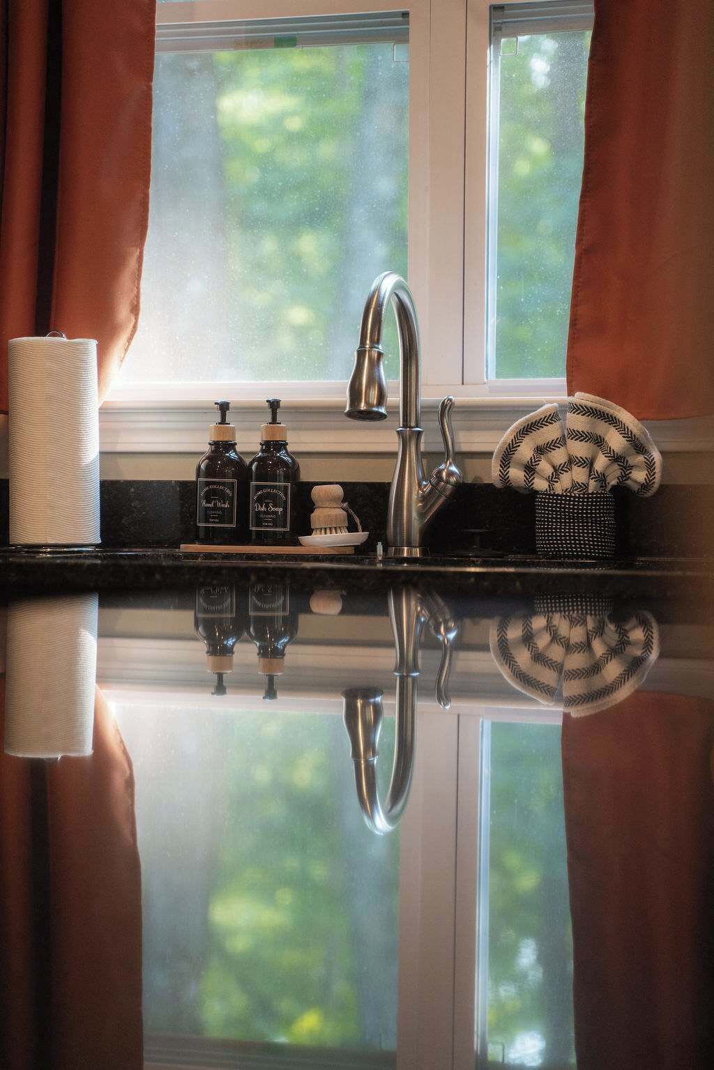 A kitchen with a stainless steel sink, a window with curtains, and a reflection on the countertop.