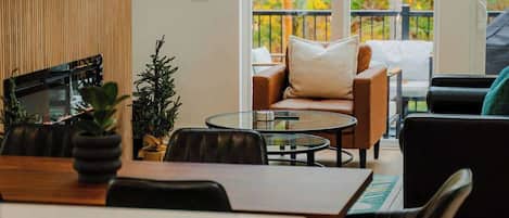 A modern dining area with a wooden table, black chairs, and a glass coffee table. There is a sofa with cushions and a view of autumn foliage through large windows.