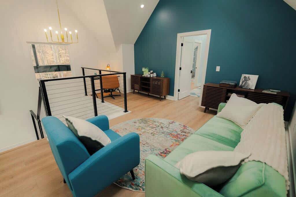 A modern living room with a high ceiling, a staircase, a green sofa, a blue armchair, and a wooden coffee table.