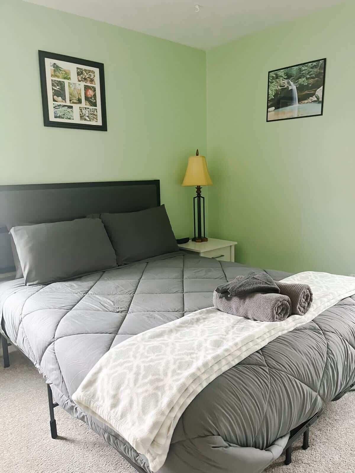 A neatly made bed with a grey quilt, a white throw blanket, and a rolled-up grey towel. There is a small bedside table with a lamp.