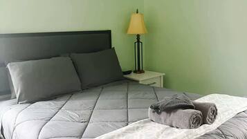 A neatly made bed with a grey quilt, a white throw blanket, and a rolled-up grey towel. There is a small bedside table with a lamp.